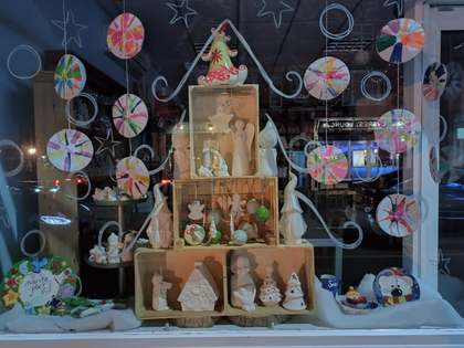 The Paint Pottle’s Christmas shop window at night, with crates of unpainted ceramic decorations stacked like a tree and hand-painted festive plates