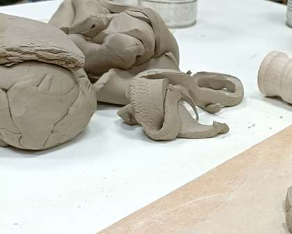 Lumps of clay and curled clay offcuts on a white workshop table, beside the end of a wooden rolling pin