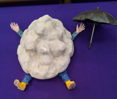 Ceramic sculpture of a figure squashed under a cloud, inscribed ‘feeling a bit under the weather’, with a tiny umbrella on a purple cloth
