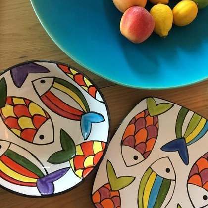 Two white dishes hand-painted with colourful fish beside a turquoise bowl of fruit on a wooden table