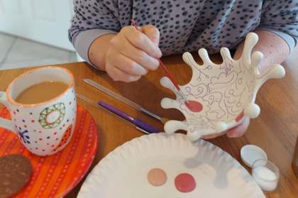 Hands painting pink dots on a white ceramic bowl, with a hand-painted mug of tea and a biscuit on the table
