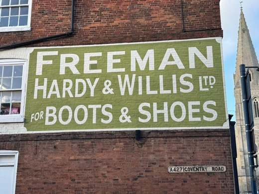 Painted ‘Freeman Hardy & Willis Ltd for Boots & Shoes’ sign on a red-brick wall above the A427 Coventry Road nameplate