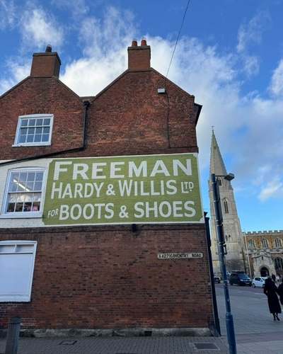Painted Freeman Hardy & Willis ‘Boots & Shoes’ sign on a brick gable, with St Dionysius church spire behind