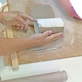 Hands shaping a slab of clay around a roll on a wooden board