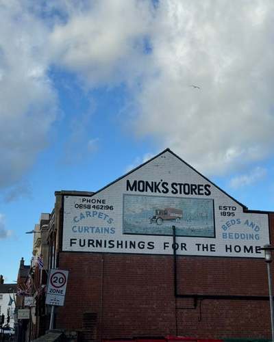 Painted Monk’s Stores sign on a brick gable advertising carpets, curtains, beds and bedding, established 1895, under a cloudy blue sky