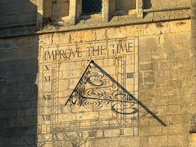 Carved sundial on the stone tower of St Dionysius Church, with the motto ‘Improve the Time’ and an ornate wrought-iron gnomon casting a long shadow.