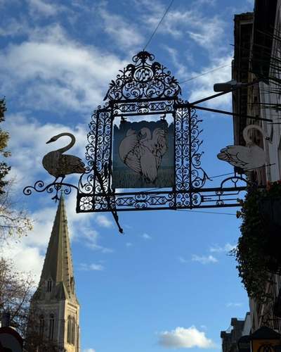 Ornate wrought-iron Three Swans hanging sign with a painted swan panel, set against a blue sky with a church spire behind