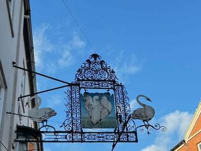 Ornate wrought-iron Three Swans sign with painted swan panel and swan silhouettes against a blue sky on the High Street