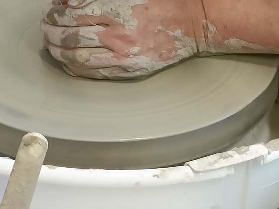 Clay-covered hands shaping clay on a spinning pottery wheel, above the splash tray