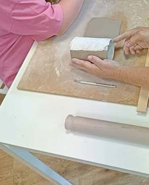 A tutor’s hands guiding a clay slab wrapped round a paper-covered former on a wooden board, beside a child’s pink sleeve